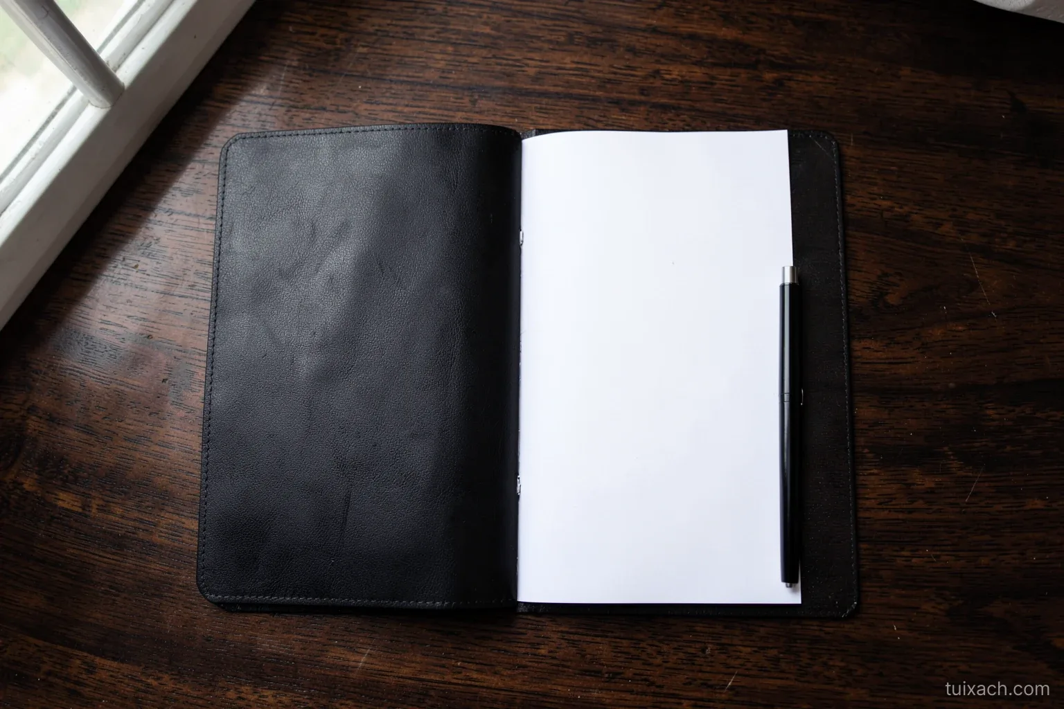 Top-down view of an open slim black leather document folio on a dark wooden desk holding blank white paper and a pen, soft window light from the left