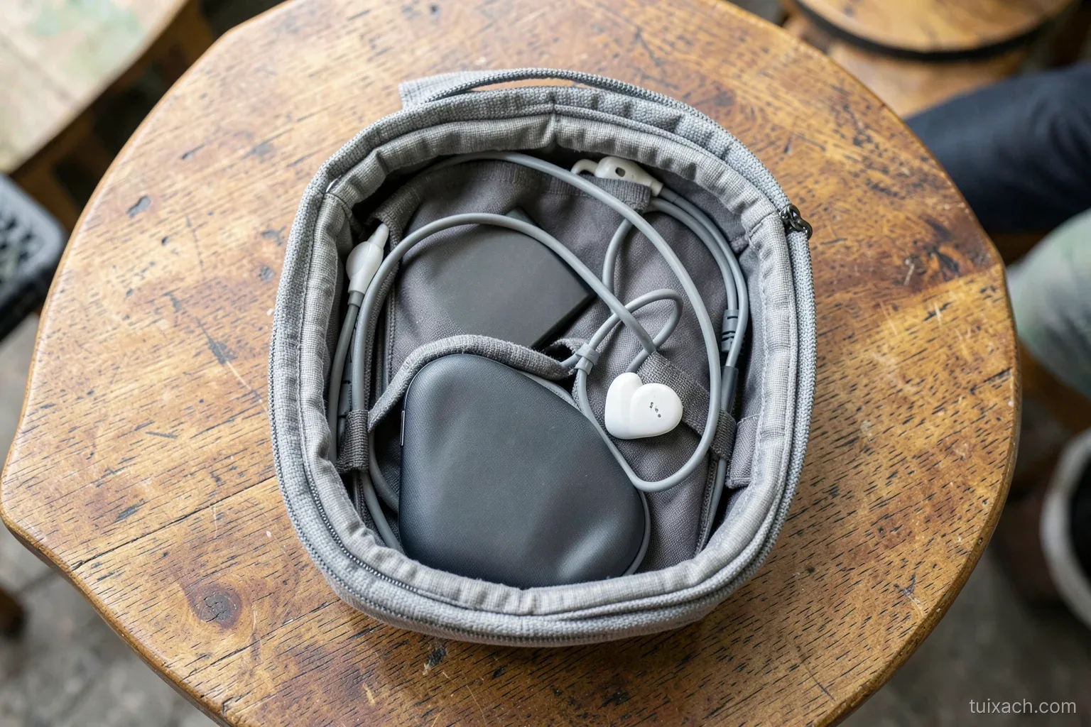 Top-down view of an open grey padded tech organizer pouch on a wooden cafe table in Da Nang, coiled cables, a power bank and earbuds case in elastic loops, soft natural light