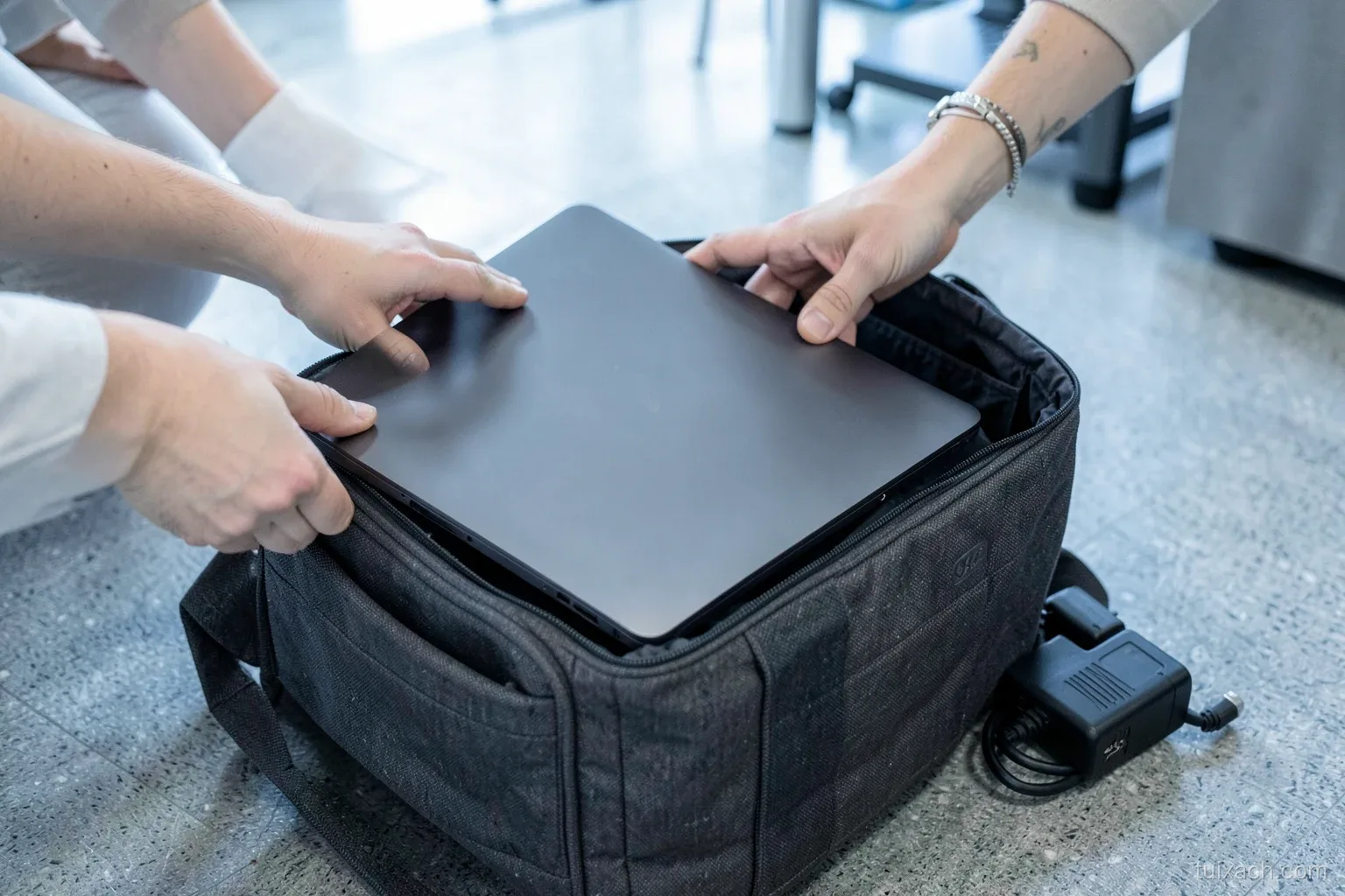 Side view of hands sliding a large closed laptop into the padded compartment of a dark grey structured bag on an office floor, bulky charger beside it, cool daylight